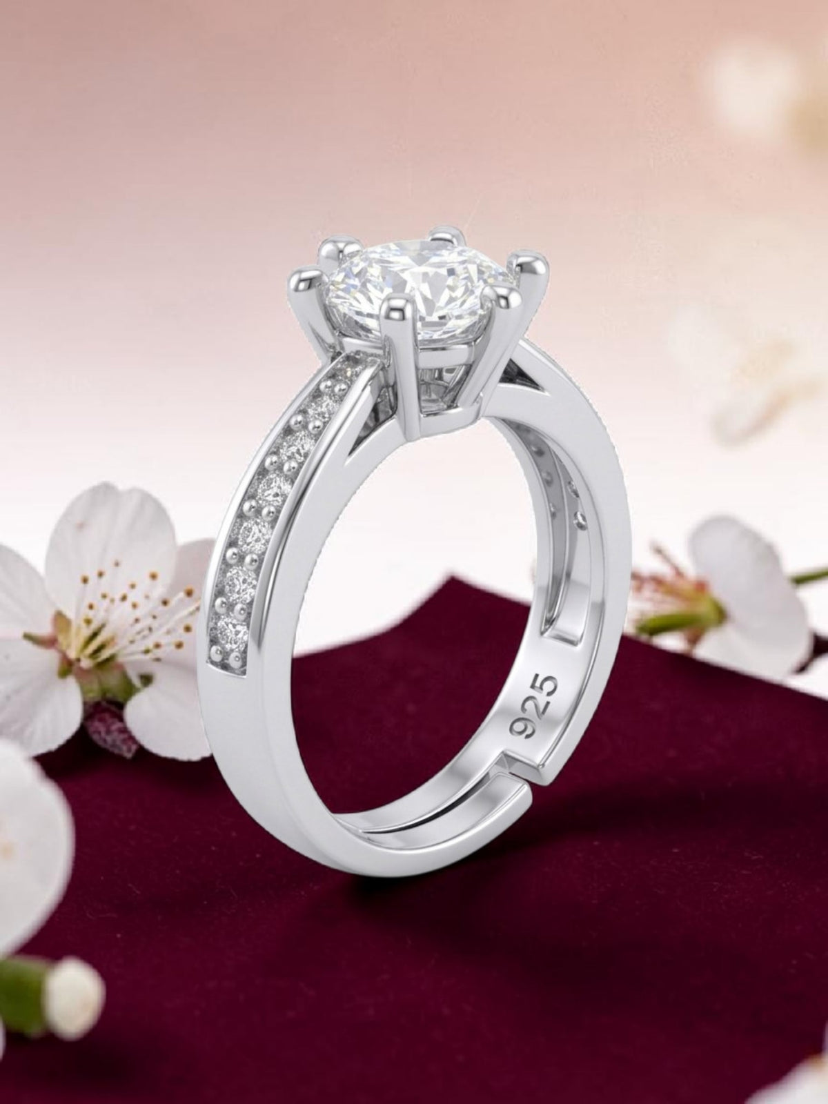 A silver-toned solitaire engagement ring with a round-cut center stone and a pavé-set accent band, displayed on a dark burgundy fabric surface with white blossoms against a soft pink background.