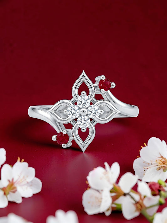 A silver-toned ring with a floral and geometric design, featuring clear stones in the center and small red gemstone accents, displayed on a deep red background with white blossoms.