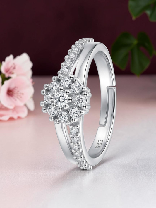 A silver-toned adjustable ring with a floral cluster of clear stones and pavé-set accents, photographed on a soft pink surface with blurred flowers and green leaves in the background.